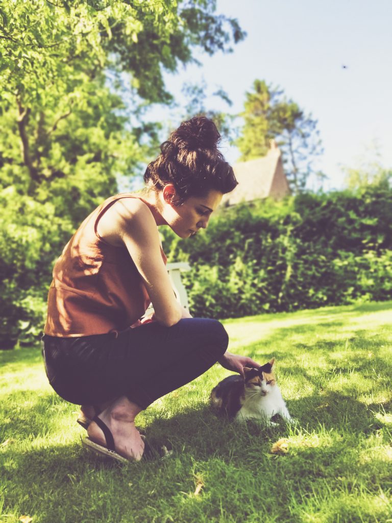 moody textile girl wearing skinny jeans and a brown shirt with black sandals in lush green leafy garden stroking a cat. Sewing Jily Top.