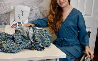 Marie Lawlor Juki Ambassador sitting in a teal wrap dress in her wheelchair in front of her Juki NX7 sewing machine on an RMF table