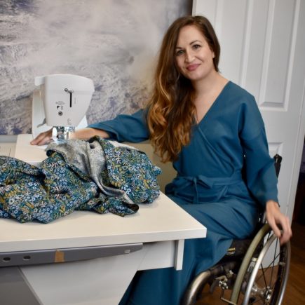 Marie Lawlor Juki Ambassador sitting in a teal wrap dress in her wheelchair in front of her Juki NX7 sewing machine on an RMF table