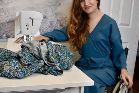 Marie Lawlor Juki Ambassador sitting in a teal wrap dress in her wheelchair in front of her Juki NX7 sewing machine on an RMF table