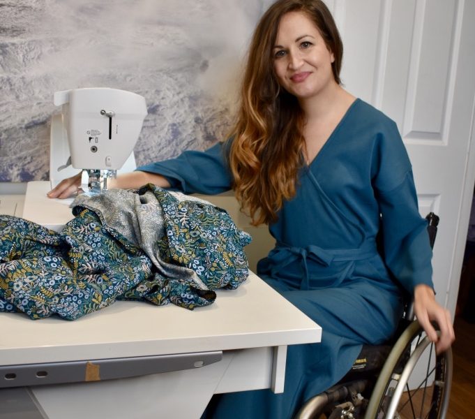 Marie Lawlor Juki Ambassador sitting in a teal wrap dress in her wheelchair in front of her Juki NX7 sewing machine on an RMF table